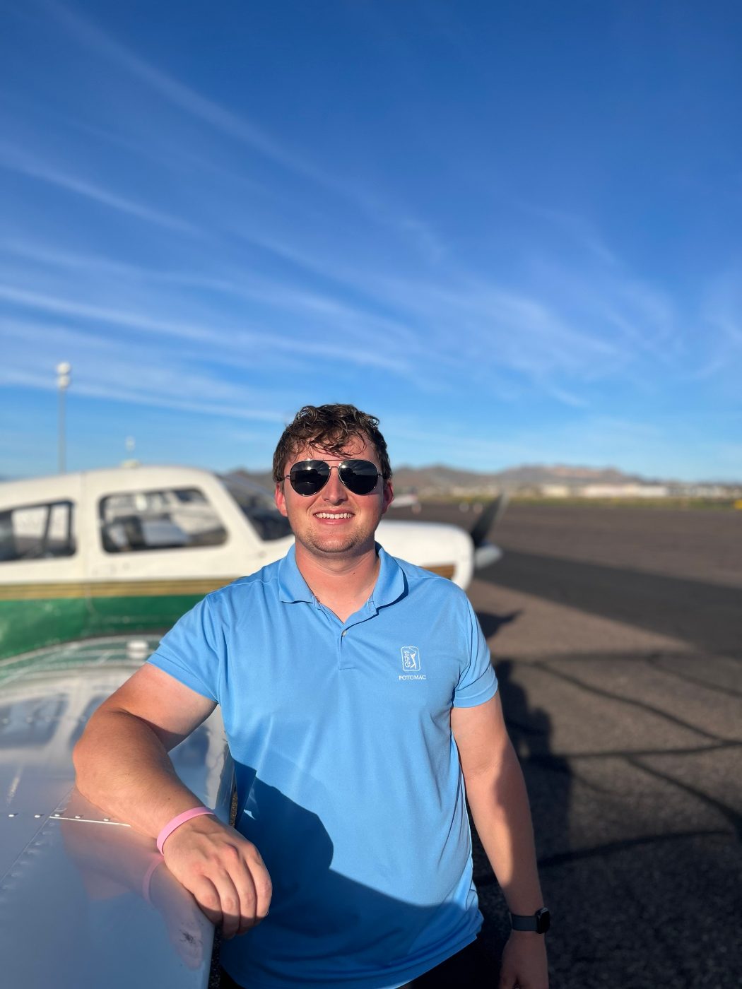 Josh Petrie, smiling in sunglasses, leaning on the wing of a Piper on the ramp under a blue sky.